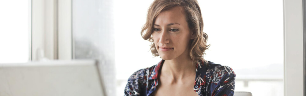 Einkommensschutz Eine Frau sitzt vor einem Laptop in einem hellen Raum. Sie hat kurze Haare und trägt eine gemusterte Bluse. Der Hintergrund ist unscharf und hell erleuchtet, was auf Tageslicht schließen lässt.